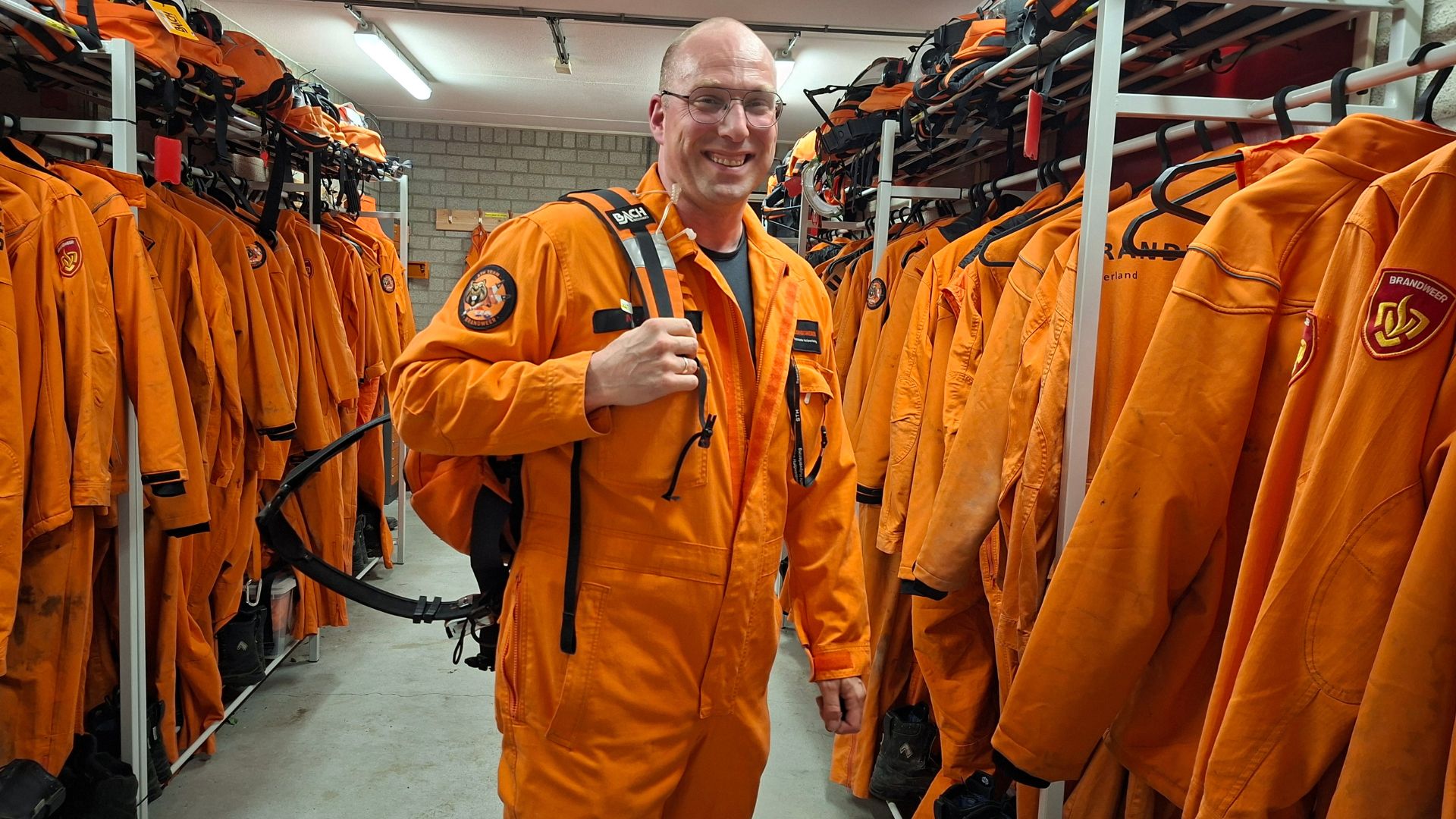 Marnix Abels staat in zijn oranje STH-overall in de kleedkamer van STH. Naast hem een lang rek met andere overalls en daarboven de helmen.